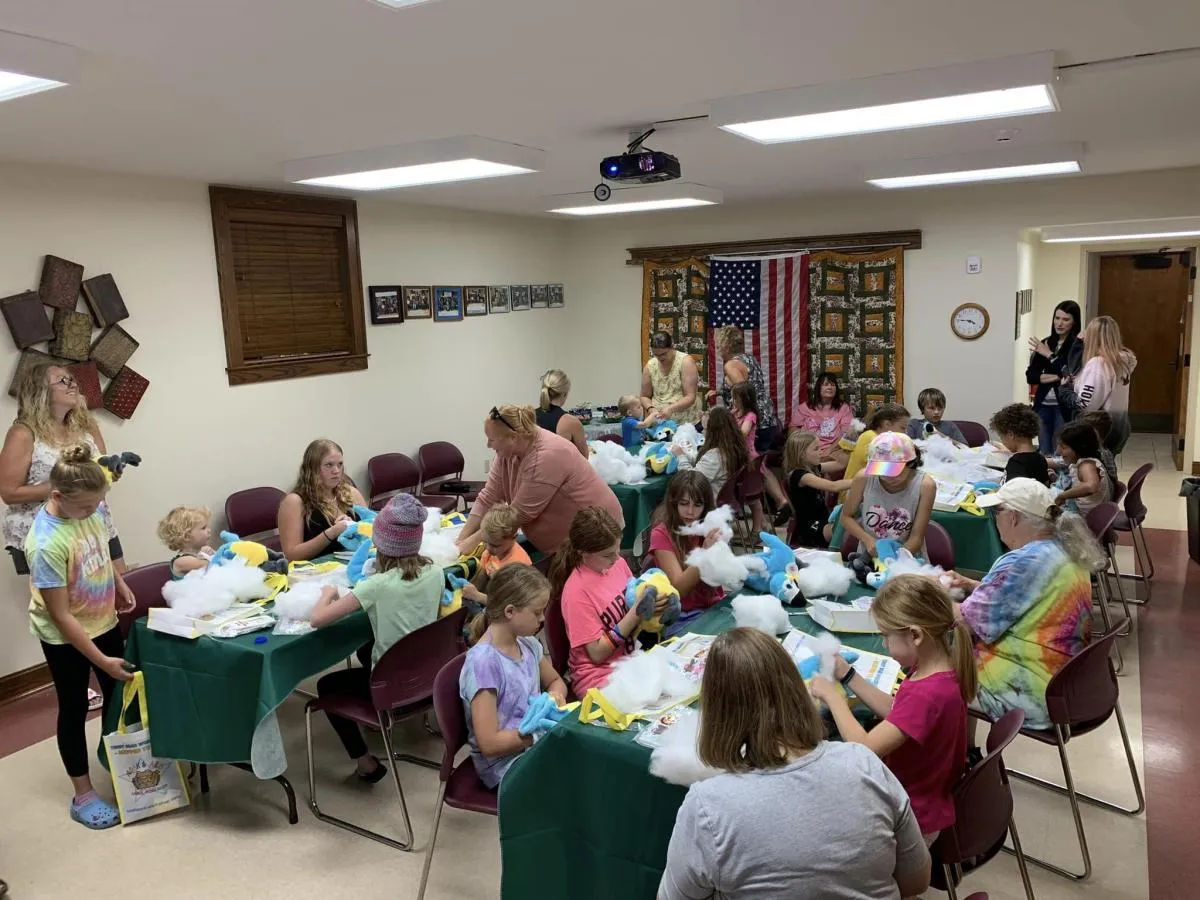 A group of children and adults are seated around tables in a room, engaged in a crafting activity.